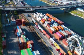 
Luftaufnahme eines Containerterminals an einem Fluss mit gestapelten Containern, Gleisen, Lkw und einer Straßenbrücke.