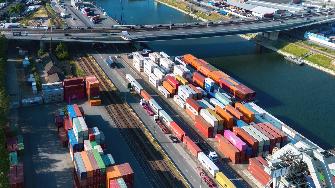 
Luftaufnahme eines Containerterminals an einem Fluss mit gestapelten Containern, Gleisen, Lkw und einer Straßenbrücke.