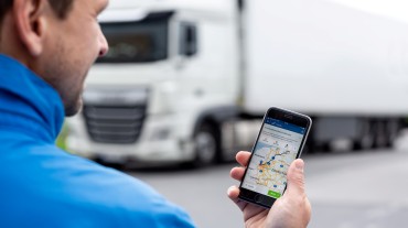 
Lkw-Fahrer hält ein Smartphone mit geöffneter Toll Collect-App und angezeigter Fahrroute in der Hand, während ein Lkw im Hintergrund parkt.
