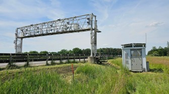 
Kontrollbrücke an einer Autobahn, daneben ein Technikhaus auf einer Wiese unter blauem Himmel