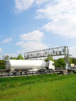 
Lkw fährt unter einer Kontrollbrücke hindurch, umgeben von grünem Gelände und Himmel im Hintergrund.