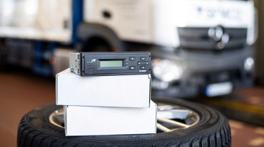 
Eine On-Board-Unit liegt auf gestapelten Kartons und einem Reifen in einer Werkstatt. Im Hintergrund ist ein unscharfer LKW zu sehen.