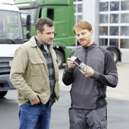 
Ein Mitarbeiter zeigt einem Mann eine On Board-Unit auf einem Betriebshof mit Lkw im Hintergrund