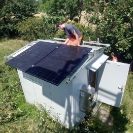 
Eine Person installiert Solarmodule auf dem Dach eines Shelters, der in einer grünen Wiese steht.