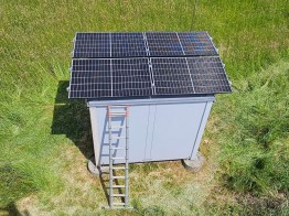 
Ein Shelter mit vier Solarmodulen auf dem Dach steht inmitten einer grünen Wiese.