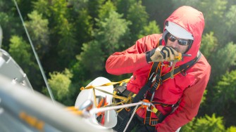 
Mann in roter Jacke hängt mit einem Seil an einem Mobilfunkmast und spricht in ein Walkie-Talkie, von oben fotografiert, im Hintergrund Bäume.