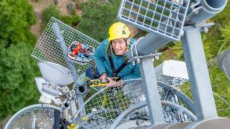 
Ein Mann mit Helm steht auf einem Mobilfunkmast und hält ein Messgerät, von oben fotografiert.