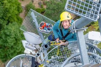 
Ein Mann mit Helm steht auf einem Mobilfunkmast und hält ein Messgerät, von oben fotografiert.