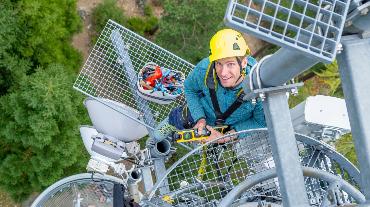 
Ein Mann mit Helm steht auf einem Mobilfunkmast und hält ein Messgerät, von oben fotografiert.