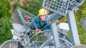 
Ein Mann mit Helm steht auf einem Mobilfunkmast und hält ein Messgerät, von oben fotografiert.