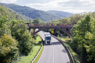 
Ein Lkw fährt auf einer Bundesstraße unter einer Brücke in einer grünen, hügeligen Landschaft.