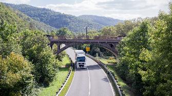 
Ein Lkw fährt auf einer Bundesstraße unter einer Brücke in einer grünen, hügeligen Landschaft.