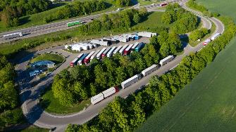 
Luftaufnahme eines von Bäumen umgebenen Lkw-Rastplatzes mit zahlreichen parkenden Lkw nahe einer Autobahn