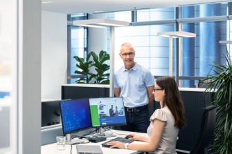 
Eine Frau arbeitet an einem Schreibtisch mit zwei Monitoren, während ein Mann neben ihr steht und mit ihr spricht.