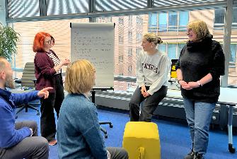 
Mehrere Personen stehen und sitzen in einem Büro vor einem Flipchart und sprechen miteinander