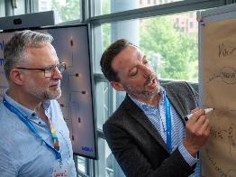 
Zwei Personen stehen vor einem Flipchart, eine schreibt darauf, während die andere zuschaut