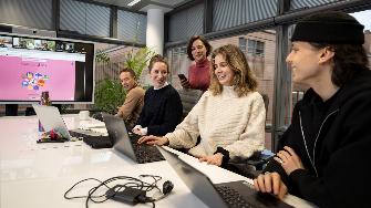 
Mitarbeiter*innen arbeiten an Laptops, während eine Videokonferenz auf einem Bildschirm im Hintergrund läuft.