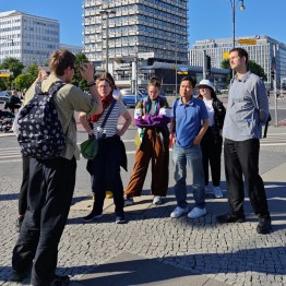 
Personengruppe hört auf einem Platz mit Hochhäusern im Hintergrund einem Mann zu, der gestikuliert.