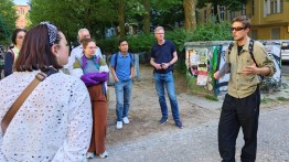 
Personengruppe hört draußen einem Mann zu, umgeben von Bäumen und Häusern.