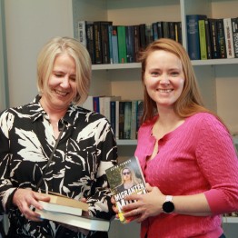 
Toll Collect Mitarbeiterinnen stehen vor Bücherregal, halten Bücher in der Hand und lächeln in die Kamera.