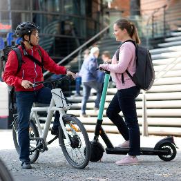
Zwei Personen unterhalten sich mit einem Fahrrad und einem E-Scooter vor einem Gebäude