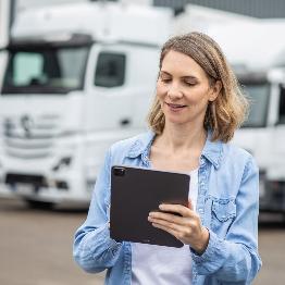 
Eine Frau betrachtet auf einem Betriebsgelände ein Tablet, im Hintergrund stehen mehrere Lkw.