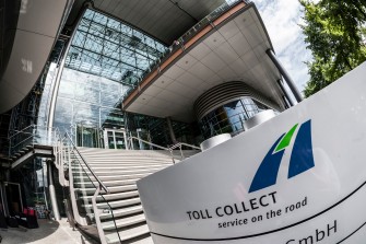 
Toll Collect Gebäude mit Glasfassade und Treppenaufgang, im Vordergrund das Firmenschild mit Logo und Slogan von Toll Collect.