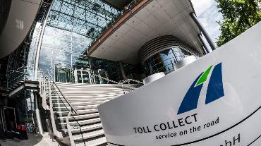 
Toll Collect Gebäude mit Glasfassade und Treppenaufgang, im Vordergrund das Firmenschild mit Logo und Slogan von Toll Collect.