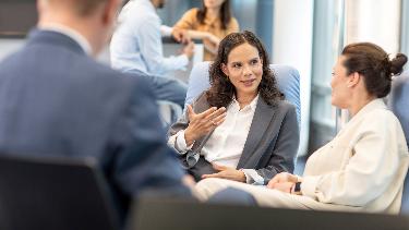 
Mehrere Personen sitzen in einem Büro zusammen; zwei Frauen im Vordergrund unterhalten sich