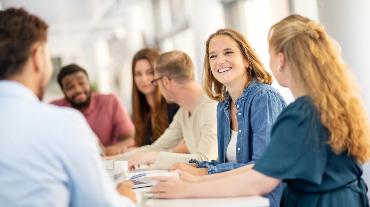 
Mehrere Personen sitzen an einem Tisch in einem Büro und unterhalten sich lächelnd miteinander.