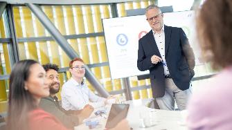 
Mehrere Personen sitzen an einem Konferenztisch, während ein Mann vor einem Bildschirm steht und präsentiert.