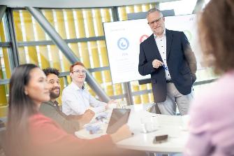 
Mehrere Personen sitzen an einem Konferenztisch, während ein Mann vor einem Bildschirm steht und präsentiert.