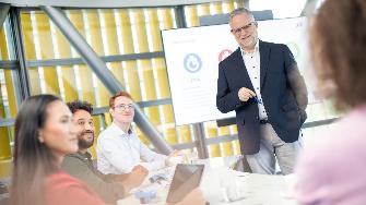 
Mehrere Personen sitzen an einem Konferenztisch, während ein Mann vor einem Bildschirm steht und präsentiert.