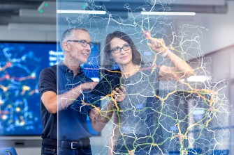 
Eine Frau und ein Mann stehen hinter einer Glasscheibe, auf der eine digitale Straßenkarte abgebildet ist.