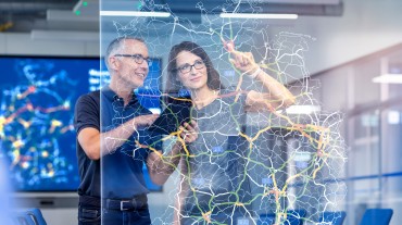 
Eine Frau und ein Mann stehen hinter einer Glasscheibe, auf der eine digitale Straßenkarte abgebildet ist.