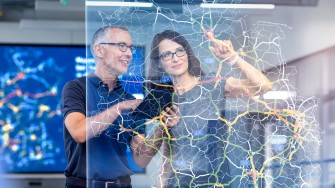 
Eine Frau und ein Mann stehen hinter einer Glasscheibe, auf der eine digitale Straßenkarte abgebildet ist.