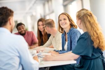
Mehrere Personen sitzen an einem Tisch in einem Büro und unterhalten sich lächelnd miteinander.