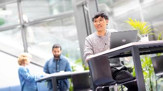 
Ein Mann sitzt an einem hohen Tisch mit einem Laptop, während im Hintergrund zwei weitere Personen arbeiten