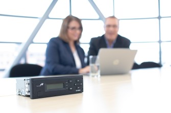 
Eine On-Board Unit von Toll Collect liegt auf einem Konferenztisch, während zwei Personen im Hintergrund am Laptop arbeiten.