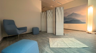 
Ruheraum mit blauen Sitzmöbeln, Trennwand, gedämpfter Beleuchtung und Wandbild mit Landschaftsmotiv.