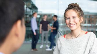 
Eine Frau lächelt in die Kamera, während im Hintergrund drei Personen im Gespräch sind.