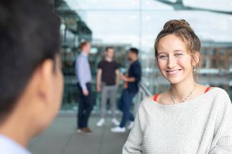 
Eine Frau lächelt in die Kamera, während im Hintergrund drei Personen im Gespräch sind.