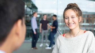
Eine Frau lächelt in die Kamera, während im Hintergrund drei Personen im Gespräch sind.