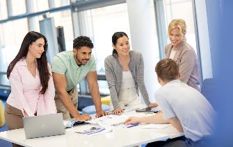 
Fünf Personen stehen um einen Tisch und besprechen gemeinsam Unterlagen.