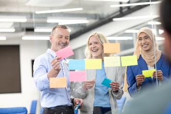 
Drei Personen stehen vor einer Glaswand mit bunten Haftnotizen und lachen, während sie Zettel in der Hand halten