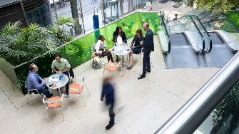 
Menschen im Gespräch im Atrium des Toll Collect Gebäudes, umgeben von Pflanzen und einer Rolltreppe.