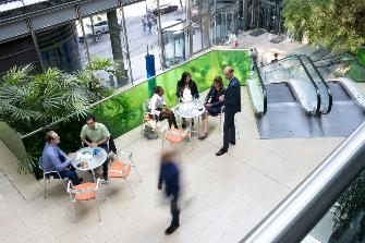 
Menschen im Gespräch im Atrium des Toll Collect Gebäudes, umgeben von Pflanzen und einer Rolltreppe.