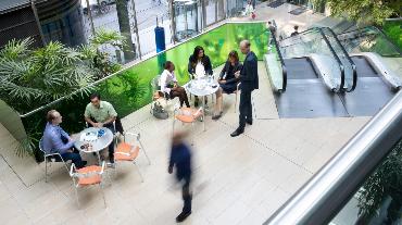 
Menschen im Gespräch im Atrium des Toll Collect Gebäudes, umgeben von Pflanzen und einer Rolltreppe.