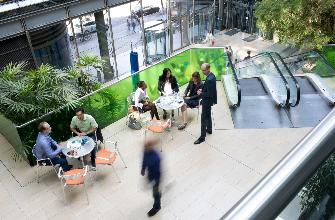 
Menschen im Gespräch im Atrium des Toll Collect Gebäudes, umgeben von Pflanzen und einer Rolltreppe.