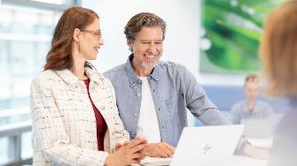 
Zwei Personen stehen nebeneinander an einem Laptop und lächeln, während sie gemeinsam auf den Bildschirm schauen.